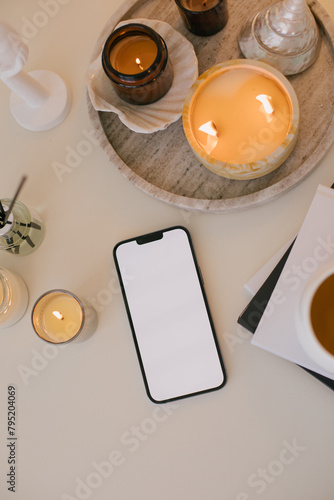 Smartphone with white empty display on a white table with candles, aromatic sticks and home decor.