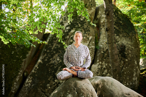 Woman practicing yoga outdoors in forest. Barefoot female on yoga mat surrounded by trees and large rocks, which suggests peaceful, natural environment ideal for meditation or yoga practice.