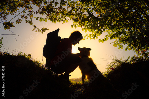 silhouette of male hiker with backpack crouching down petting and cuddling his border collie dog in a forest at sunset
