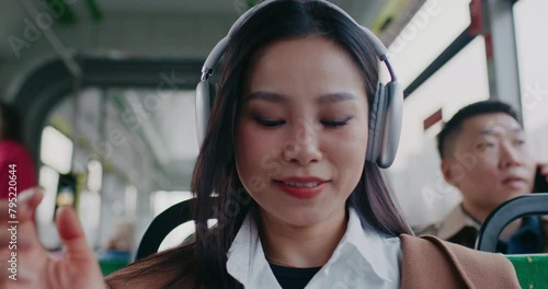 Facial portrait of Asian girl commuting in public transport. Passengers in blurred background. Woman putting on large new wireless headphones to enjoy good quality music. Using technology gadgets.