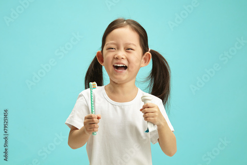 Cheerful little asian ethnicity girl holding squeezed toothpaste tube and toothbrush, fun laughing, wearing in white t shirt, standing over blue isolated background.