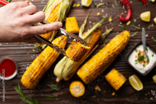 Obraz na płótnie Kitchen tongs with grilled corn on the cob pieces in caucasian hands on kitchen wooden table background w salonie