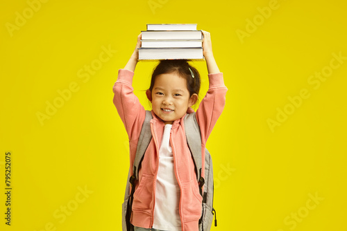 Sweet first grader asian ethnicity schoolgirl holding books above head, cheerful smiling standing with backpack on her shoulders over yellow isolated background.