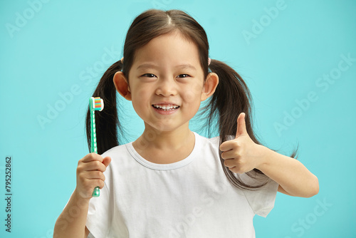 Sweet chinese child kidl wearing in white t shirt use toothbrush, showing thumb up gesture, stands over blue isolated background with a free copy space. Children oral hygiene concept.