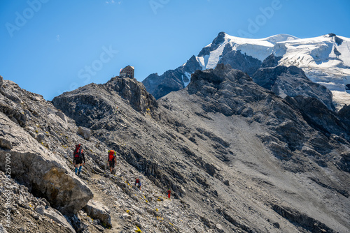 A group of hikers trek along a rocky trail with the snow-capped peak of Ortles mountain towering in the background under a clear blue sky.