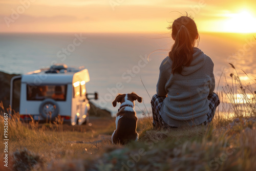 A woman and her dog sit near a campervan by the sea.