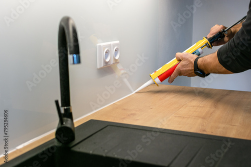 Glazier is using a silicone caulking gun to fill gap between glass backsplash and countertop in new modern kitchen