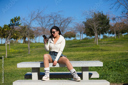Young and beautiful Spanish brunette woman with sunglasses sitting on a park bench. The girl is casually dressed and consulting her mobile phone while enjoying the sunny spring day.