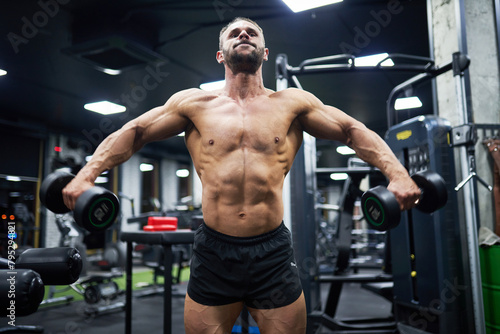 Shirtless Caucasian bodybuilder working out chest muscles  while using dumbbells. Low angle view of male crossfit athlete exercising in modern gym at evening. Concept of sport  bodybuilding.