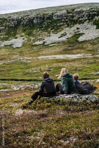 Mother and two sons enjoying a rest while hiking in a nordic landscape. A family takes a break on a rock during a hike, with the mother pointing out something in the distance. © Dylan