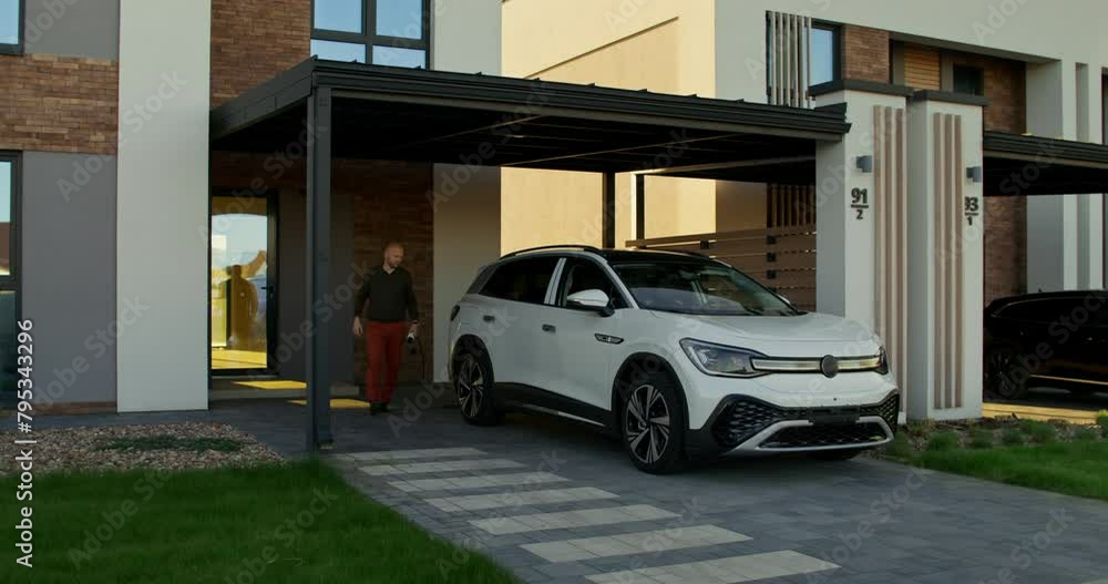 A man in casual clothes connects a charger to an electric car parked in front of a stylish cottage. A sunny day A man in casual clothes connects a charger to an electric car parked in front of a stylish cottage. A sunny day