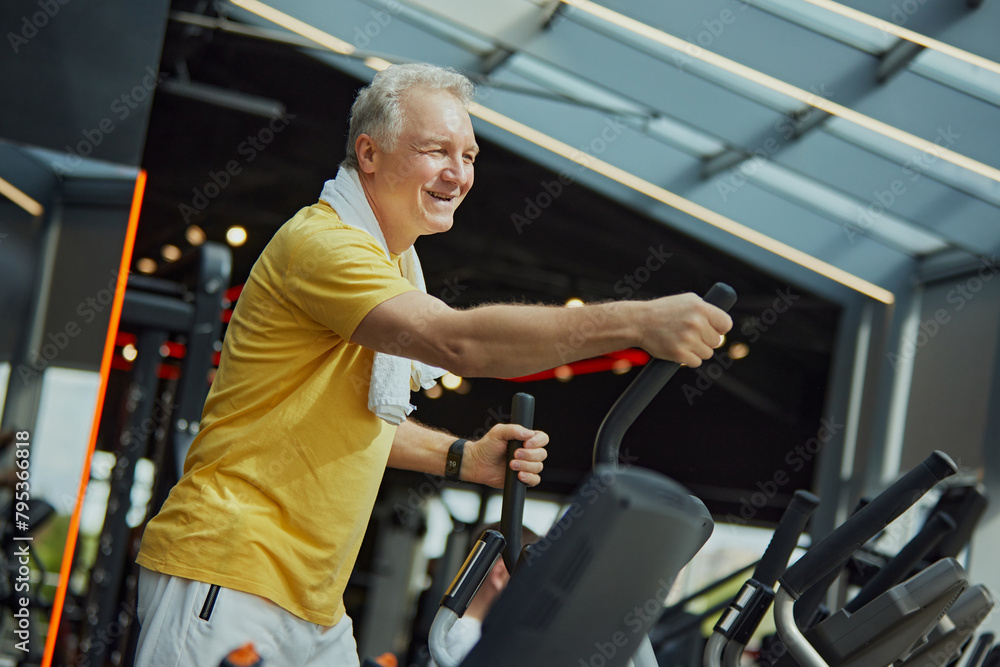 Positive, smiling mature man in vibrant sports attire with towel on shoulders work out on exercise machine at modern gym. Concept of sport, active seniors in modern life, healthy lifestyle. Ad Positive, smiling mature man in vibrant sports attire with towel on shoulders work out on exercise machine at modern gym. Concept of sport, active seniors in modern life, healthy lifestyle. Ad