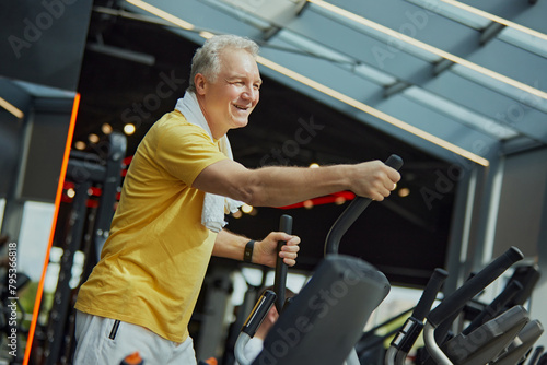 Positive, smiling mature man in vibrant sports attire with towel on shoulders work out on exercise machine at modern gym. Concept of sport, active seniors in modern life, healthy lifestyle. Ad