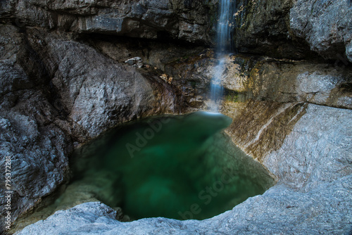 pozza di acqua verde smeraldo e trasparente nella penombra, che si è formata in una conca rocciosa, in fondo ad una piccola cascata di montagna photo