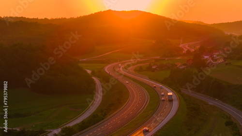 AERIAL, LENS FLARE: Morning rays peek over hill and reflect from driving vehicles. Sun rises over a highway winding through hilly countryside, casting long shadows and painting sky in orange shades.