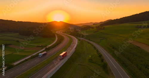 AERIAL, LENS FLARE: A stunning sunset over hilly landscape and winding highway. Calm evening and light traffic on the motorway passing green countryside colored in orange hue of setting summer sun.