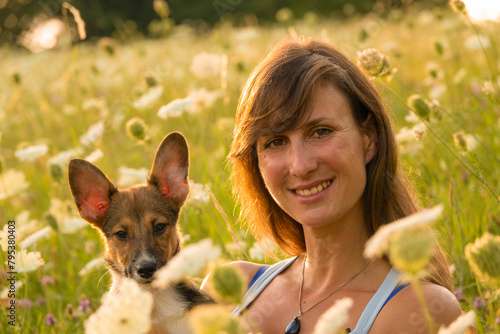 PORTRAIT, DOF: Cheerful lady poses with her young adopted puppy amidst lush blooming wildflowers. Woman holds her doggo as they sit surrounded by the beauty of a blossoming meadow in golden sunlight. © helivideo