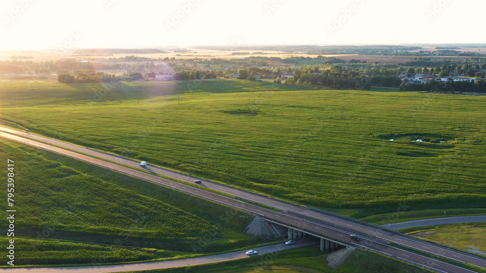 Vehicles driving on asphalt multi lane expressway. Intercity road passes through rural corn green fields. Journey travel cars drive on highway with overpasses and bridges at sunset. Aerial view