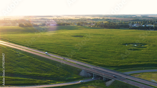 Vehicles driving on asphalt multi lane expressway. Intercity road passes through rural corn green fields. Journey travel cars drive on highway with overpasses and bridges at sunset. Aerial view
