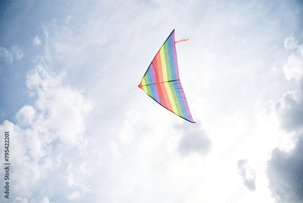 Colorful rainbow Kite flying high in the blue gray sky. Isolated kite on the blue background. Weekend leisure entertainment activities. Peaceful relaxation. Time with loved one, children and family. Colorful rainbow Kite flying high in the blue gray sky. Isolated kite on the blue background. Weekend leisure entertainment activities. Peaceful relaxation. Time with loved one, children and family.