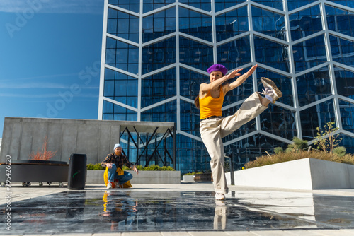 Vibrant Outdoor Scene Captures A Young Woman In A Yellow Top And Purple Hat Dancing Energetically, With A Male Spectator In Yellow Pants And A Blue Hat, Against A Modern Glass Building Background.