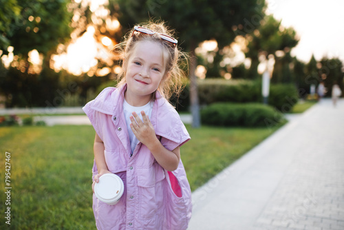 Cute stylish child girl 5-6 year old holding coffee cup and wearing sun glasses outdoor over sun light in park. Looking at camera. Summer season