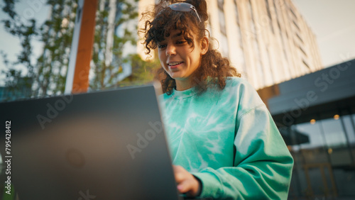 Young Woman With Curly Hair, Wearing A Turquoise Sweatshirt, Engaging With A Laptop Outdoors In An Urban Setting, Showcasing A Blend Of Technology And Casual Lifestyle.