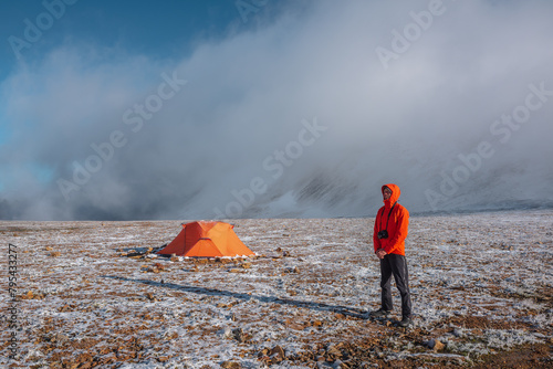 Man in red jacket in hood with photo camera near orange tent on snow-covered stony pass in sunlight at misty early morning. Freshly fallen snow in foggy high mountains at sunrise. Guy in low clouds.