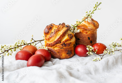 Freshly baked pastries next to red eggs and delicate white flowers on textured fabric. Easter holiday concept.