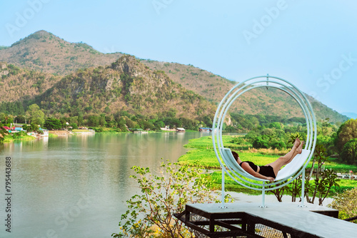 Happiness woman relaxing on comfortable seat with beautiful view of nature at balcony on hill near riverbank. Female resting on curved seats on hillside terrace with mountains and river in background.