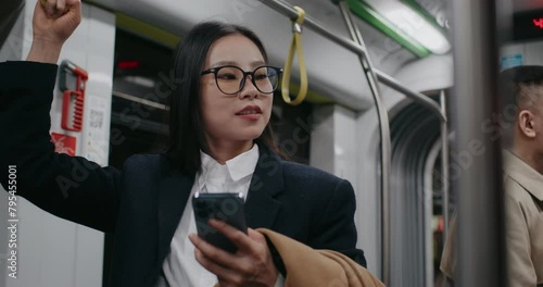 Frontal view of stylish Chinese girl holding on to safety handle inside moving bus or train. Checking on news or new posts in her smartphone. Beginning to jiggle with joy after reading something funny