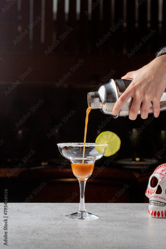 A professional bartender skillfully pours a vibrant red cocktail into a martini glass, garnished with a slice of lime, against a dark bar background