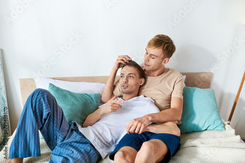 A man in casual attire lays next to another man on a bed, sharing a moment of intimacy.