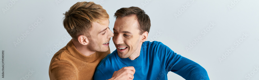 Two men, a loving gay couple, in casual attire, standing next to each other on a gray backdrop. Two men, a loving gay couple, in casual attire, standing next to each other on a gray backdrop.
