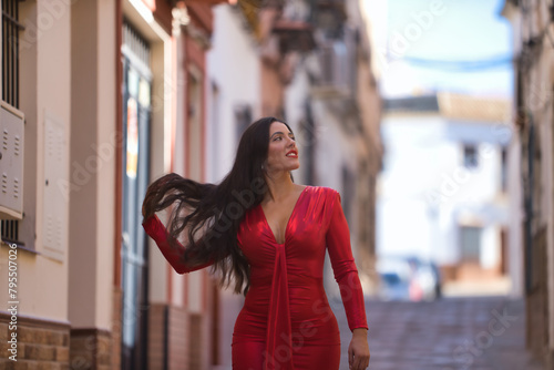 Young, beautiful brunette woman in an elegant red dress, walking while touching her hair, at dawn down a lonely street of a beautiful white Andalusian village. Concept beauty, fashion, trend, travel. © Manuel