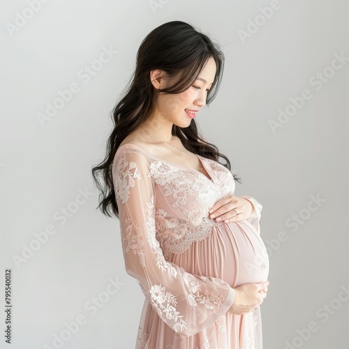 Pregnant asian woman in light pink thin dress happily rubs her belly, woman side view, white background 