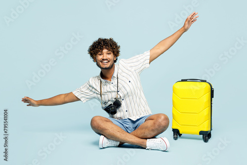 Full body traveler Indian man wear white casual clothes sit near bag with outstretched hands isolated on plain blue background. Tourist travel abroad in free time rest getaway Air flight trip concept #795541004