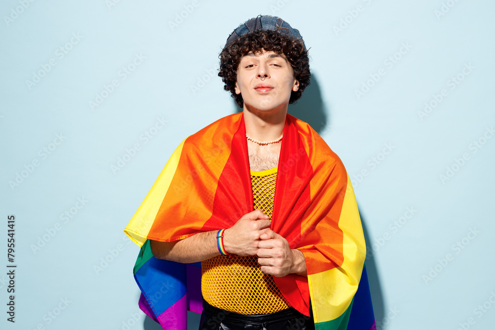 Young happy gay Latin man wears mesh tank top hat clothes wrapped in striped rainbow flag isolated on plain pastel light blue cyan background studio portrait. Pride day June month love LGBT concept.