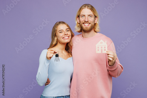 Young smiling couple two friends family man woman wearing pink blue casual clothes together hold bunch of keys and wooden house mockup isolated on pastel plain light purple background studio portrait.