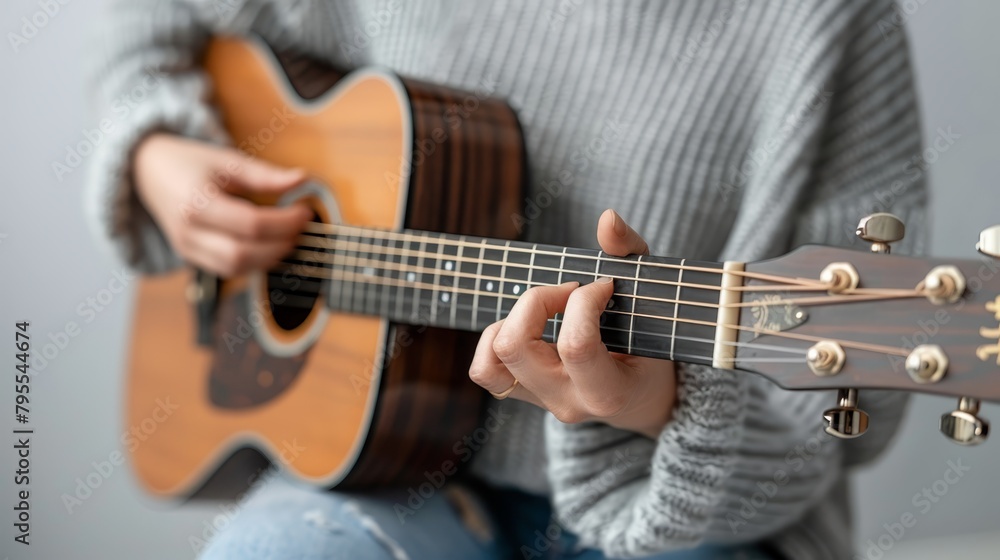   A person closely holds a guitar and plays the ukulele against a gray backdrop In the foreground, another person also holds a ukulele