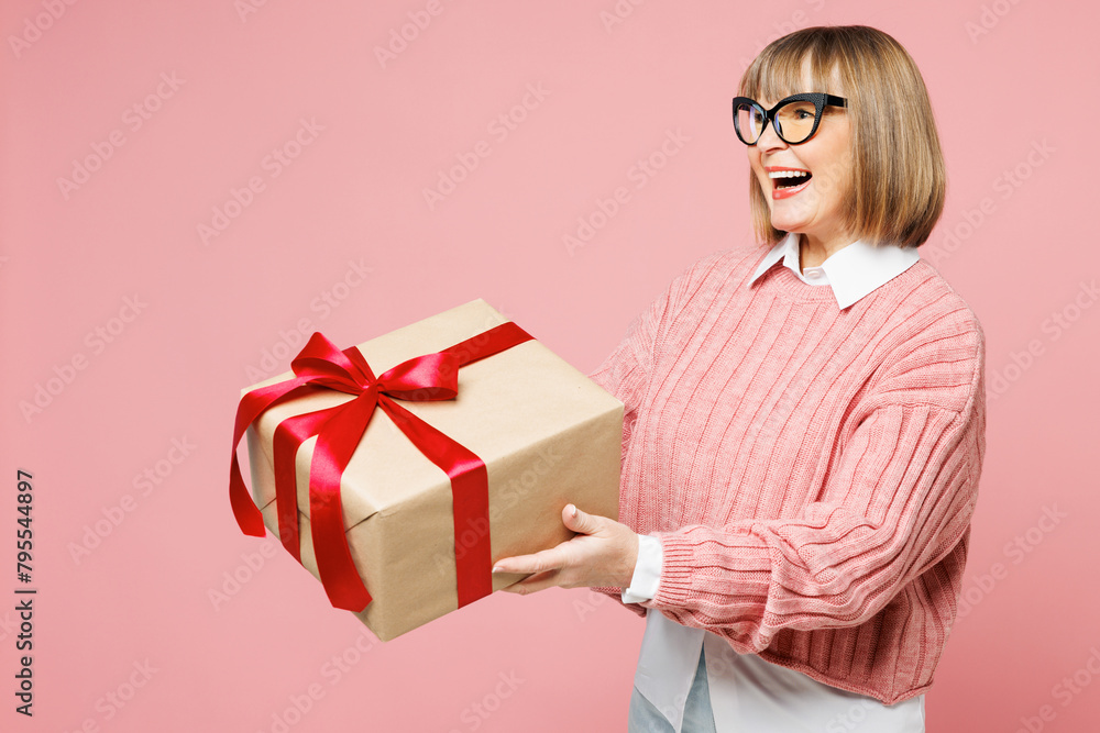 Side view elderly smiling woman 50s years old wear sweater shirt casual clothes glasses hold give present box with gift ribbon bow isolated on plain pastel pink background studio. Lifestyle concept. Side view elderly smiling woman 50s years old wear sweater shirt casual clothes glasses hold give present box with gift ribbon bow isolated on plain pastel pink background studio. Lifestyle concept.