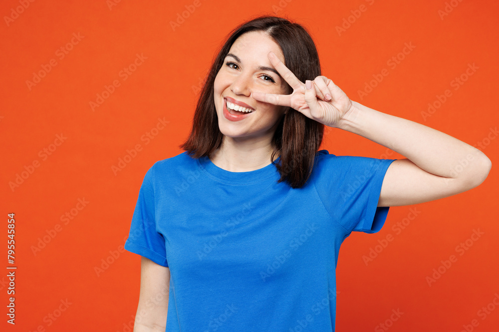 Fototapeta premium Young satisfied cheerful smiling fun happy woman she wear blue t-shirt casual clothes cover eye with v-sign looking camera isolated on plain red orange background studio portrait. Lifestyle concept.