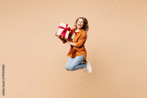 Full body young excited woman she wear orange shirt casual clothes jump high hold present box with gift ribbon bow isolated on plain pastel light beige background studio portrait. Lifestyle concept. photo