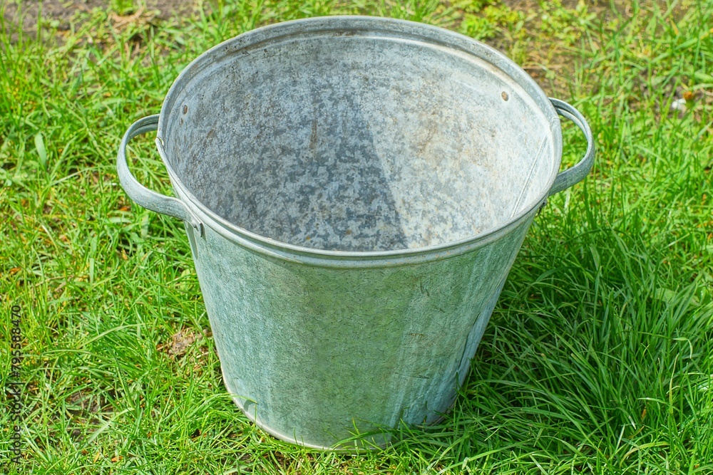 one iron gray round industrial with two handles old empty bucket stands on the green grass during the day on the street one iron gray round industrial with two handles old empty bucket stands on the green grass during the day on the street