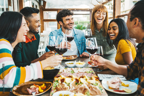 Happy friends drinking red wine sitting at restaurant table - Multiracial young people enjoying rooftop dinner party together - Food and beverage concept with guys and girls having lunch break outside photo