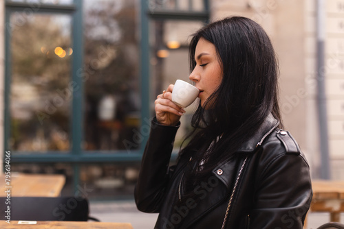 Thoughtful brunette teen woman drinking coffee in cafe outdoor, lifestyle, coffee, communication and local business, unaltered. Sensual woman sitting side. Girl wear leather jacker and look pretty. photo