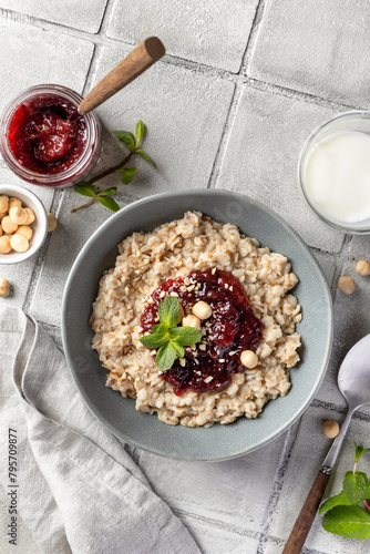 Oatmeal porridge with raspberry jam and nuts in gray bowl on gray tile background served with spoon, top view, text space. Healthy diet breakfast
