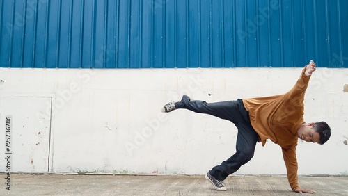 Hispanic man stretch arms and dance street dancing in front of wall. Motion shot of stylish dancer or choreographer in casual outfit practicing dancing in hip hop style. Outdoor sport 2024. Endeavor.