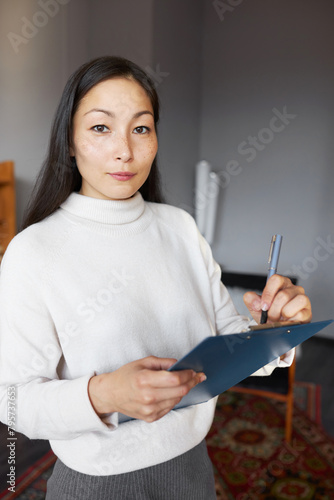 Indoor portrait of pretty female designer standing with clipboard in hands, examining living-room to make new project, making notes, looking at camera with confident facial expression