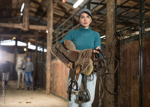 Portrait of young horsewoman walking in stable with saddle in hands, about to harness horse for riding